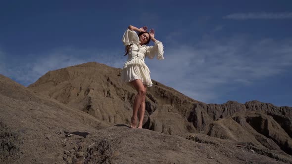 a Young Attractive Slender Woman in an Unusual White Dress Poses Standing on the Sandy-gray Hills.