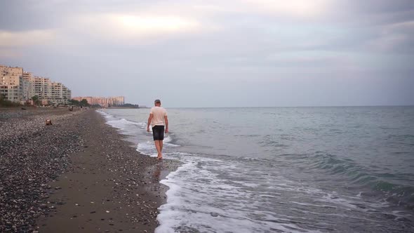 A Slender Elderly Grayhaired Man Slowly Walks Barefoot on a Deserted Beach in the Running Waves in