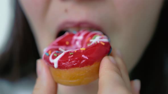 A Woman Pleasantly Smiling Bites a Soft Donut Covered with Red Fruit Glaze and Confectionery