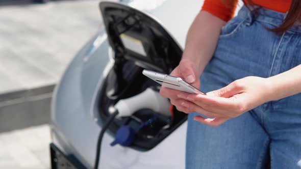 Attractive Woman Uses Phone While Charging Electric Car Near the Shopping Center