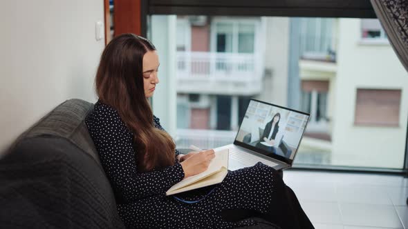 Young Focused Woman Sitting on Sofa at Home Making Notes During Distant Chat with Psychologist