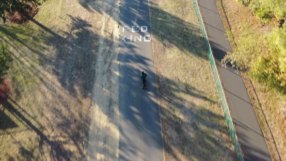 An aerial tracking of a man on an electric skateboard in an empty park on a sunny day. The drone fol