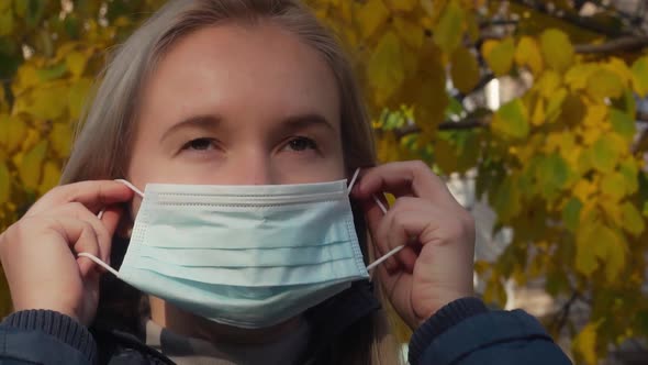 Young Woman Puts Medical Mask on Her Face on Sunny Autumn City Street. Personal Protective Measures