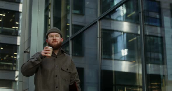 A Young Bearded Man Walks Near an Office Building and Drinks Coffee