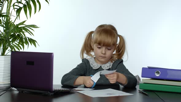 Child Schoolgirl Learns Lessons at Home Sitting at Table Cutting with Scissors Shapes Out of Paper