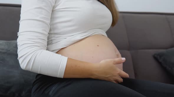 A Pregnant Fairskinned Woman is Sitting on a Cozy Sofa and Stroking Her Tummy
