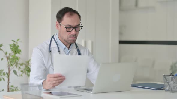 Middle Aged Doctor with Laptop Reading Documents in Office