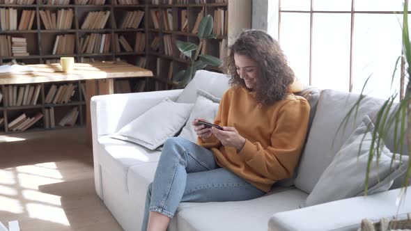 Smiling Hispanic Girl Sit on Sofa Hold Phone Watching Funny Social Media Video