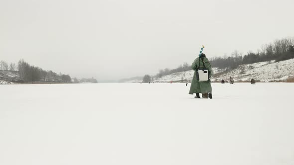 Fisherman with ice drill and fishing equipment is walking along frozen ice lake