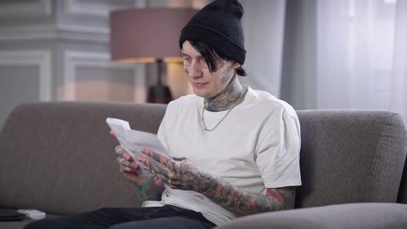 Positive Tattooed Man in Casual Clothes and Hat Putting Letter in Envelope