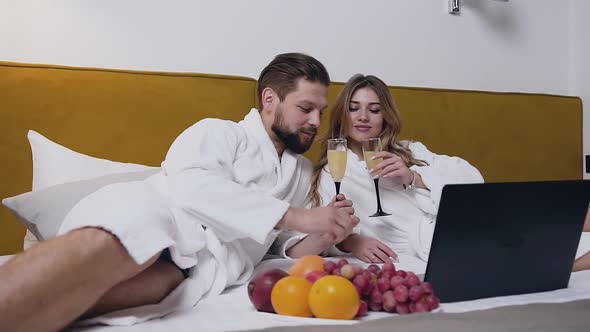 Young Couple in white Bathrobes which Lying on Bed and Drinking