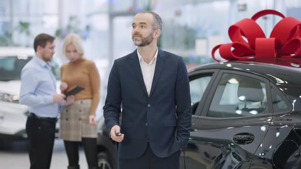 Satisfied Caucasian Businessman Standing with Car Keys at Black Luxurious Vehicle in Showroom with