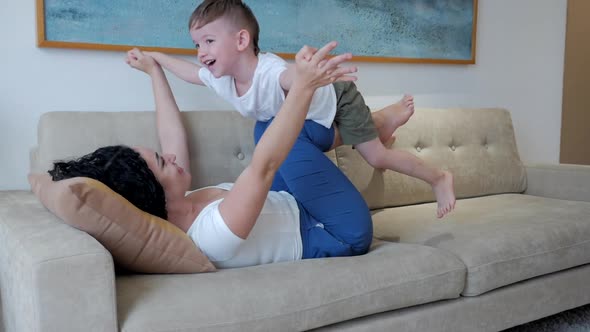 Happy Healthy Family Young Mom Lifting Cute Little Child Son Up Playing Plane on Sofa at Home, Funny