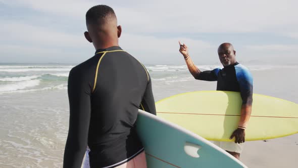 African american father and teenage son standing on a beach holding surfboards and talking