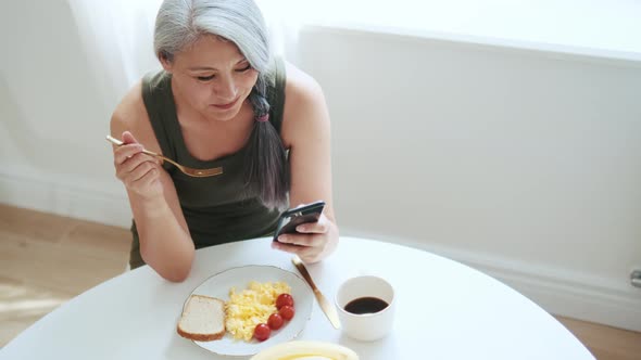 Asian smiling woman eating her breakfast and looking at the phone