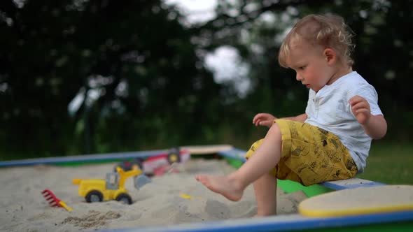 Kid in Yellow Shorts and a White Tshirt Sits in a Sandbox and Plays with Plastic Toys