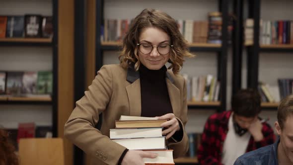 Slow Motion Footage of a Girl in Brown Jacket and Glasses Walking By Row with Pile of Books She Took