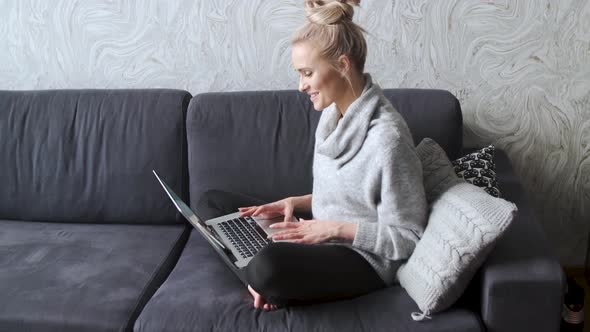 Cheerful Young Blond Woman Sitting on Couch in Living Room and Using Laptop