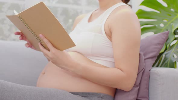 Happy Pregnant Woman sitting on couch holding book and reading for prepare to take care baby