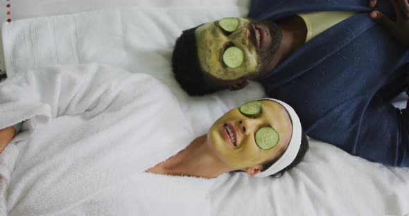 Happy diverse couple lying with masks and pieces of cucumber on eyes in bathroom
