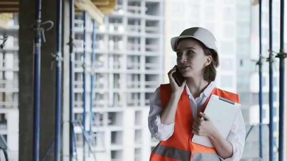 Young female civil engineer in safety jacket and helmet is talking on mobile phone