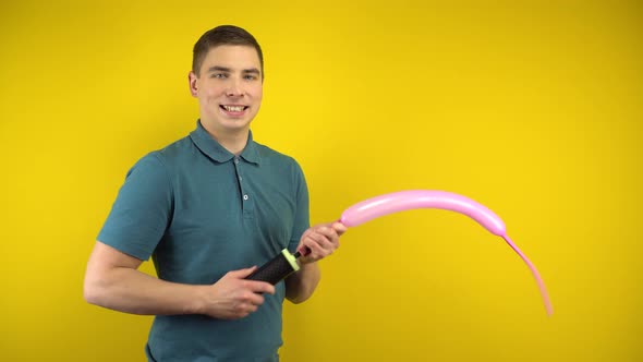 A Young Man Inflates a Long Pink Balloon with a Pump on a Yellow Background. Man in a Green Polo.