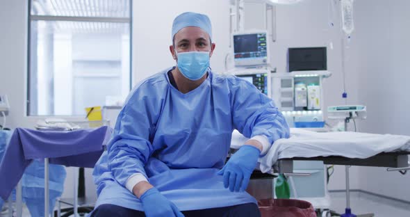 Portrait of caucasian male surgeon wearing face mask sitting in operating theatre at hospital