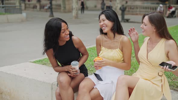 Two Young Multiethnic Women Greeting Female Friend Outdoors While Sitting in City with Coffee to Go