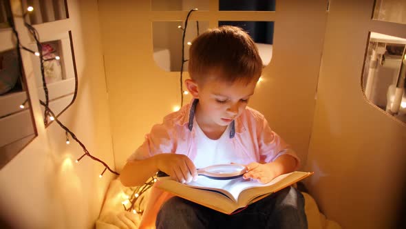 Portrait of Little Boy Holding Glashlight and Magnifying Glass While Reading Book at Night