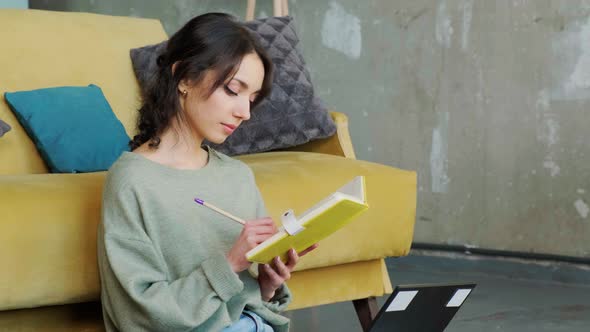 Young Girl is Browsing at Her Laptop and Writing Notice on Her Paper Notebook