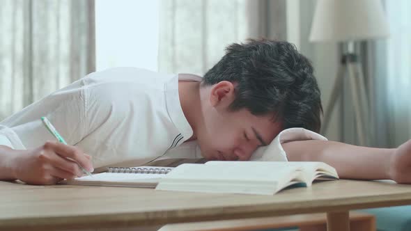 Close Up Of Asian Man Student Being Sleepy While Writing In The Notebook On The Table At Home