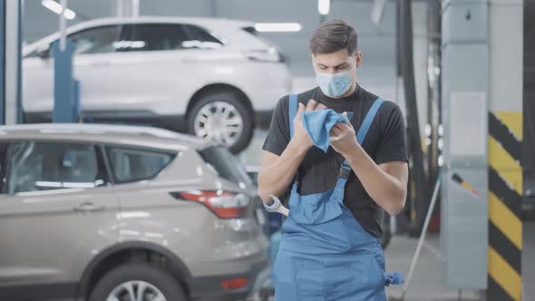 Tired Young Caucasian Auto Mechanic in Covid Face Mask Cleaning Hands in Auto Repair Shop