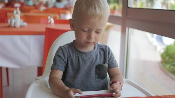 Cute Little Boy Sitting at Street Cafe Looking at the Phone. Pretty Child Sitting in the Cafe