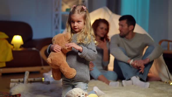 Wide Shot Portrait of Serious Pretty Girl Playing with Teddy Bear As Blurred Father and Mother