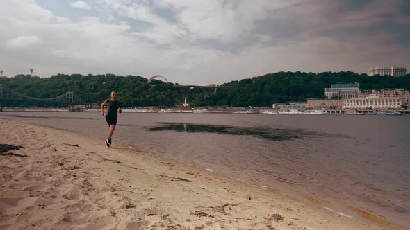 Muscular Young Man Running on the Beach in the Town on a Sunny Day