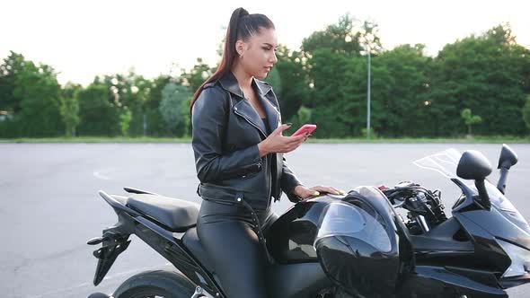 Woman Sitting on Attractive Black Motorcycle on Sportground and Using her Phone