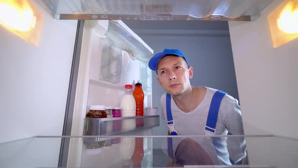 repairman looks into refrigerator, puts his hands on his head and is surprised