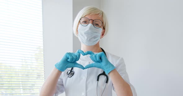 Smiling woman doctor in a medical coat, mask and stethoscope showing hands heart shape. 