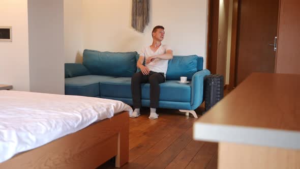 Wide Shot Portrait of Confident Young Caucasian Man Sitting on Couch in Hotel Room with Coffee Cup