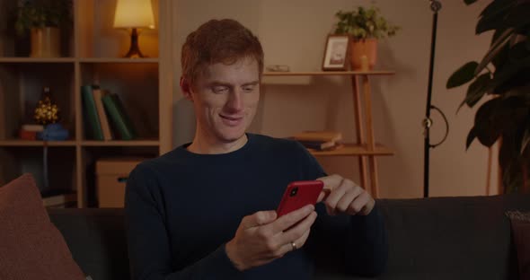Crop View of Positive Red Haired Man Using and Scrolling Smartphone Screen While Sitting on Sofa at