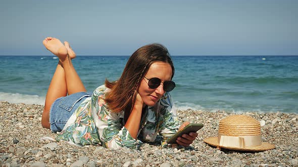 Charming hipster girl with brown hair in summer tropical shirt and jeans shorts lying on beach