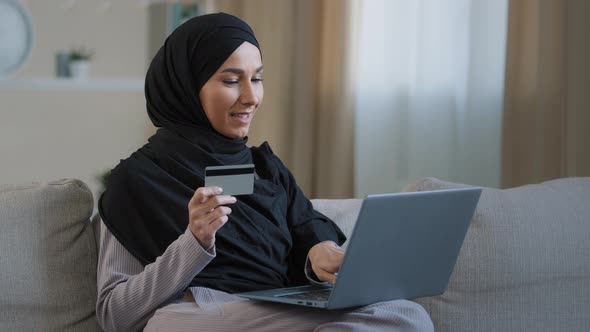 Smiling Islamic Girl Customer in Hijab Sitting on Sofa Make Online Order Use Laptop Enter Credit