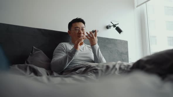 Positive Asian man wearing eyeglasses recording voice message by mobile