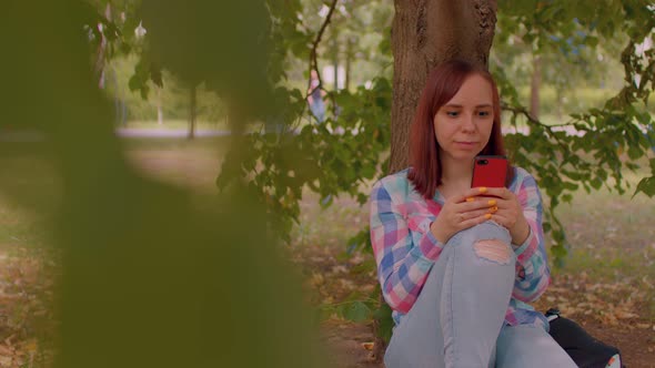 Close Up of Young Woman Rests with Mobile Phone Sitting Near Tree in Park