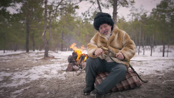 Wide Shot Focused Bearded Old Indigenous Man Sitting in Winter Forest with Fire Burning at