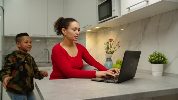 Boy Preventing Busy Mother From Work at Home Covering Eyes with Hands