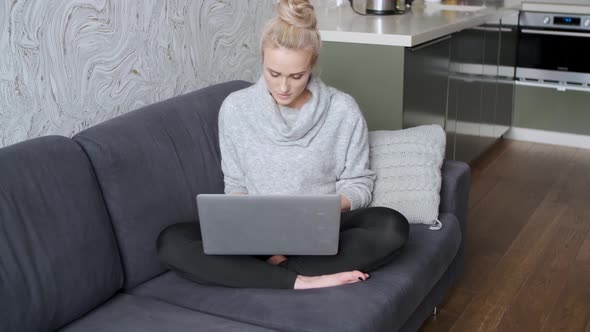 Cheerful Young Blond Woman Sitting on Couch in Living Room and Using Laptop