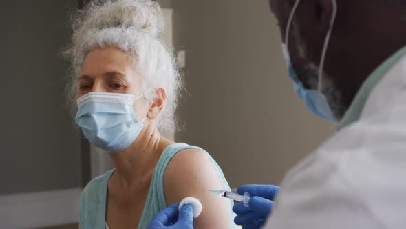 Male african american doctor wearing face mask injecting covid-19 vaccine into senior female patient