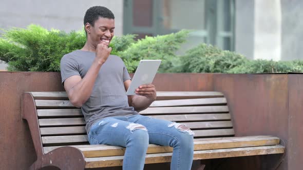 Young African Man Celebrating Success on Tablet on Bench