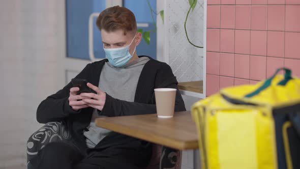 Young Caucasian Man in Covid Face Mask Surfing Internet on Smartphone Waiting for Take Away Order in
